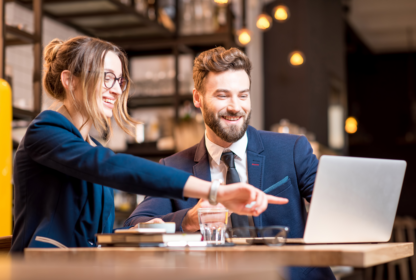 Two business professionals in a cafe setting working together on a laptop, smiling and discussing something on the screen. The woman is pointing at the laptop while the man looks on.