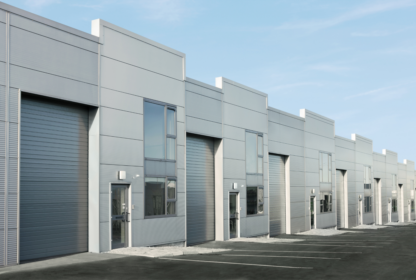 A row of modern industrial warehouse units with large roller doors and adjacent windows, set against a clear sky, with an empty parking lot in the foreground.