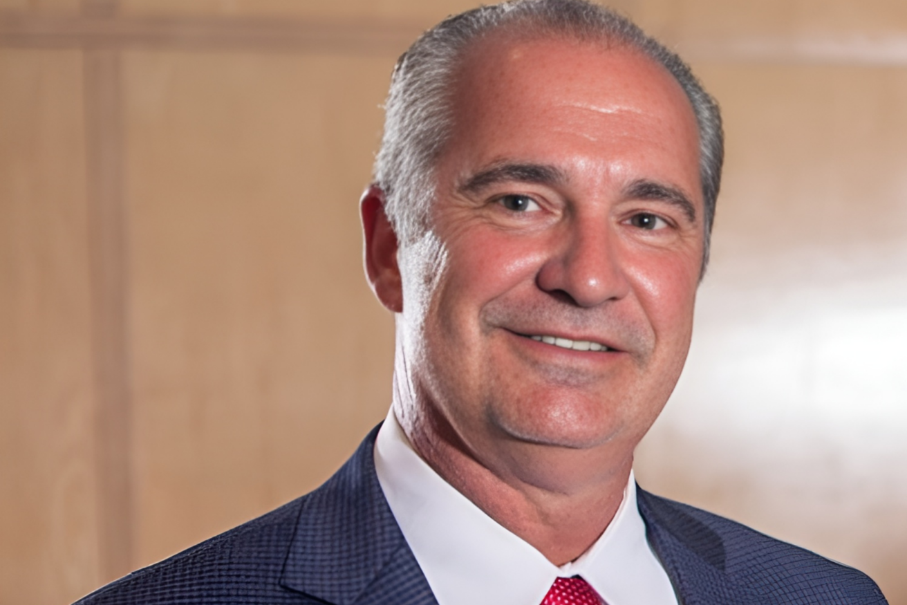 A middle-aged man with gray hair wearing a blue suit, white shirt, and red tie, smiling in front of a wooden background.