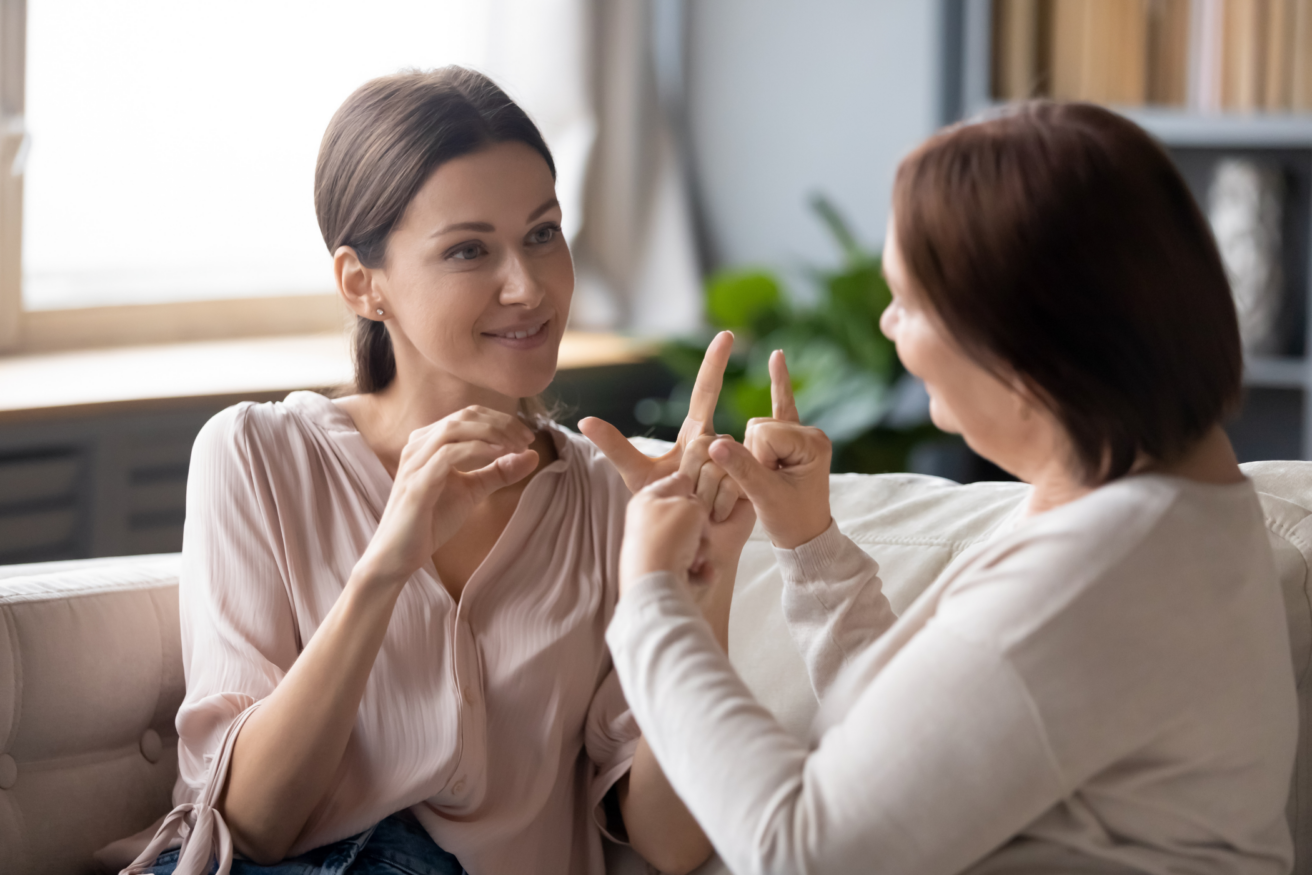 Two women sitting on a couch engage in a conversation using sign language. The younger woman faces the camera, while the older woman faces her. Bookshelves and a plant are visible in the background.