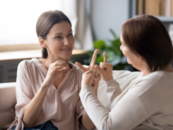 Two women sitting on a couch engage in a conversation using sign language. The younger woman faces the camera, while the older woman faces her. Bookshelves and a plant are visible in the background.