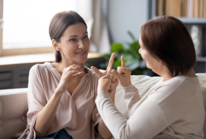 Two women sitting on a couch engage in a conversation using sign language. The younger woman faces the camera, while the older woman faces her. Bookshelves and a plant are visible in the background.