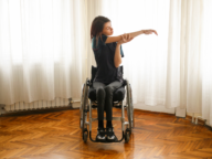 A person in a wheelchair performs a stretching exercise with one arm extended sideways and the other arm holding the elbow, in a room with wooden floors and large windows with white curtains.