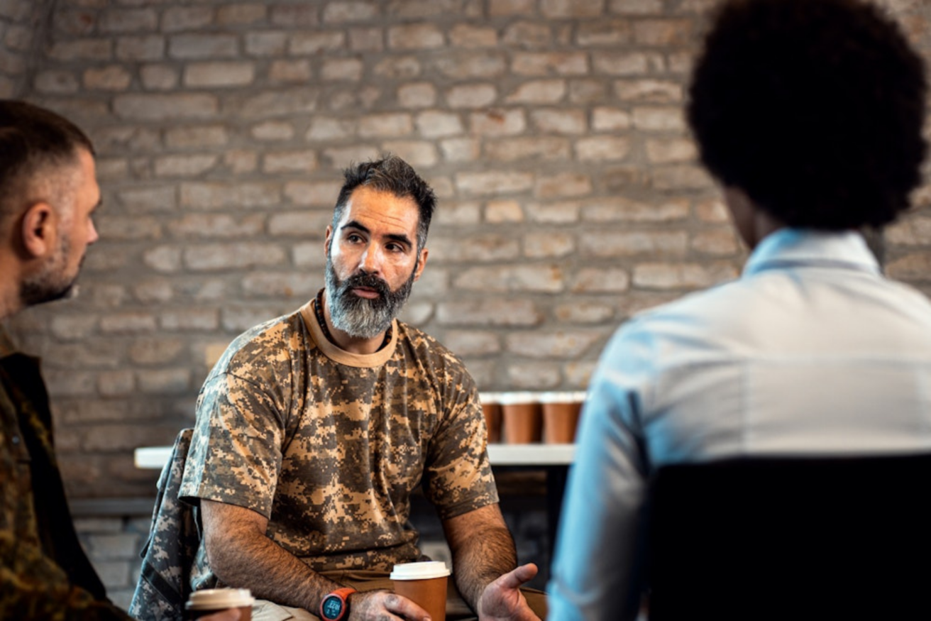 A man in a camouflage shirt is talking to two individuals in front of a brick wall. He holds a paper cup while engaged in conversation.