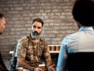 A man in a camouflage shirt is talking to two individuals in front of a brick wall. He holds a paper cup while engaged in conversation.