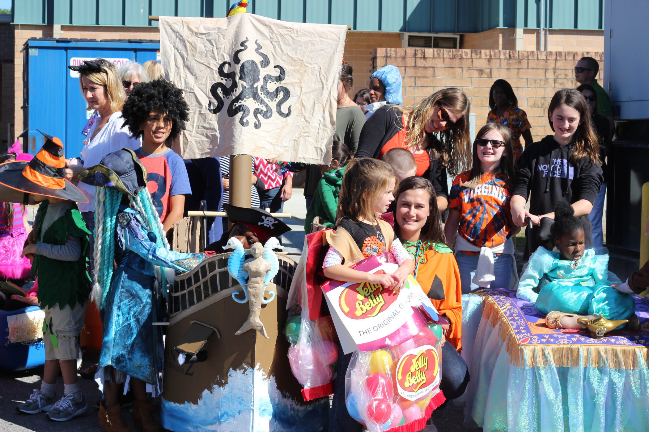 Group of people, including children in costumes, gathered outdoors by a painted pirate ship prop and holding signs at an event.