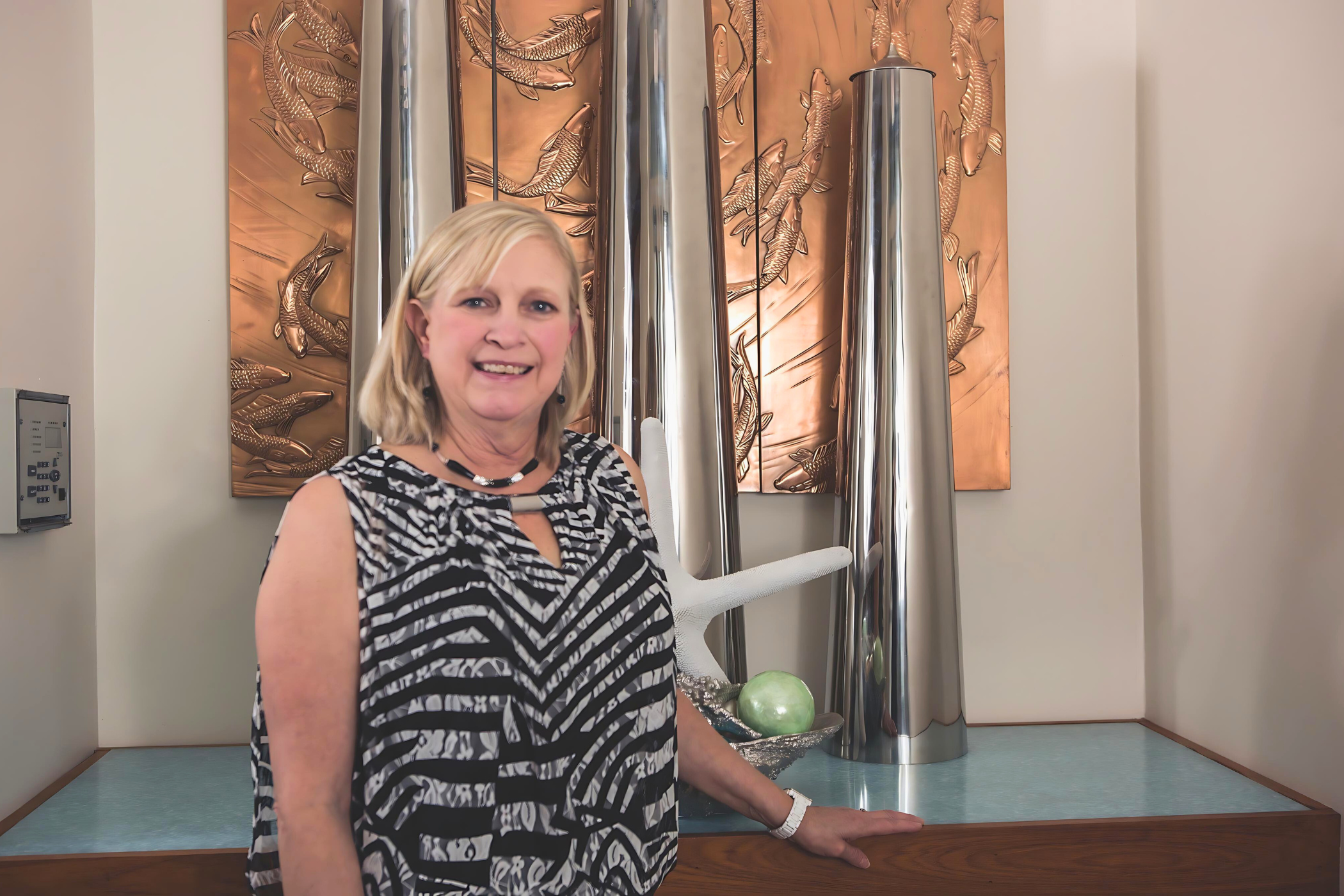 A woman with blonde hair stands in front of a decorative background featuring large cylindrical metal sculptures and a carved panel. She is wearing a black and white patterned sleeveless top.