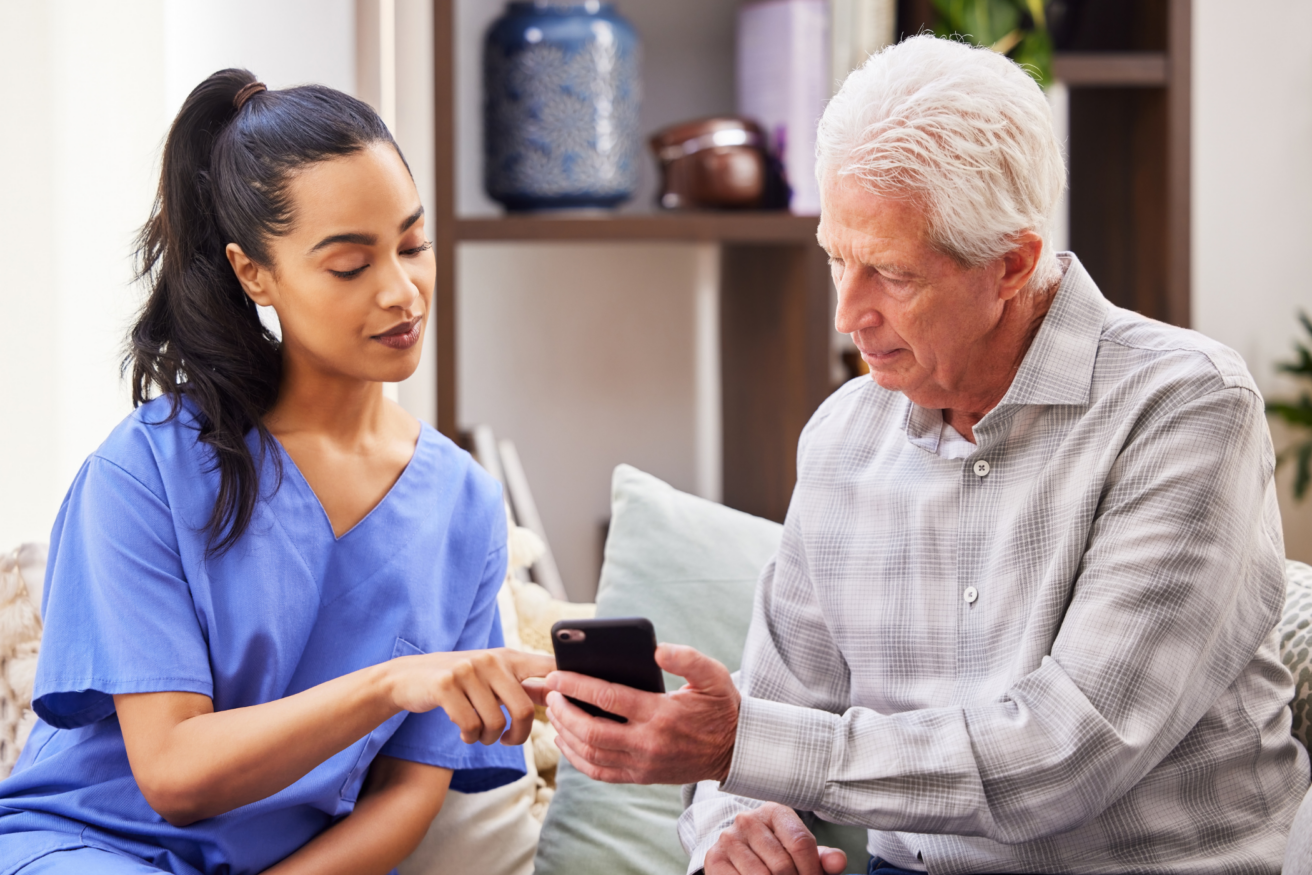 A healthcare worker in blue scrubs helps an elderly man use a smartphone while sitting on a couch.