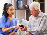 A healthcare worker in blue scrubs helps an elderly man use a smartphone while sitting on a couch.