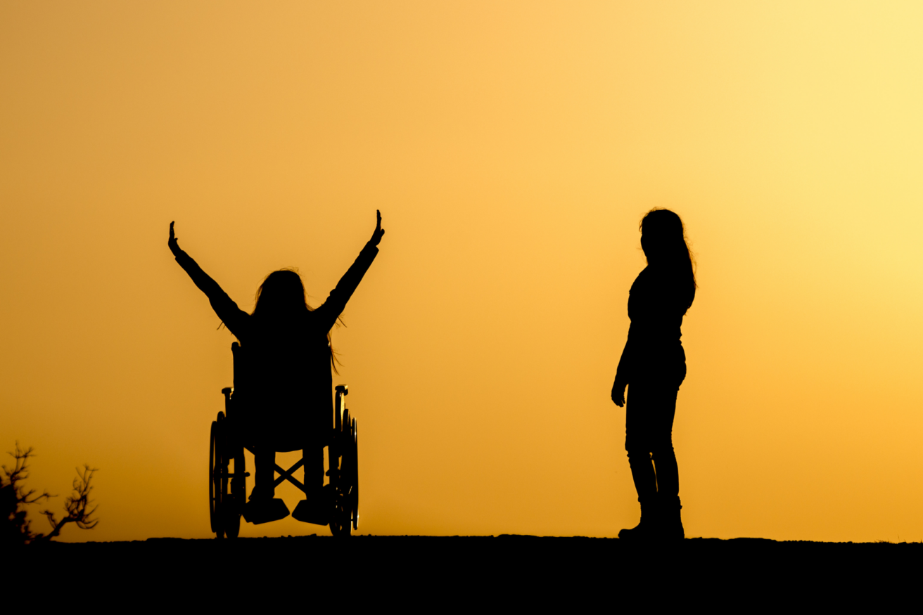 Silhouetted against a sunset, a person in a wheelchair raises their arms in celebration while another person stands nearby.