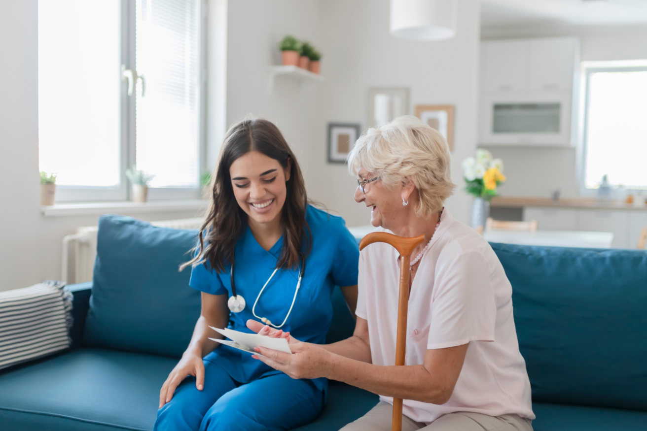 A smiling healthcare worker in blue scrubs sits on a couch with an elderly woman holding a cane, both looking at a document.