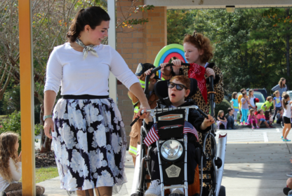 A woman in a floral skirt walks outside with a child in a wheelchair and another child standing behind. People wearing colorful outfits are in the background.