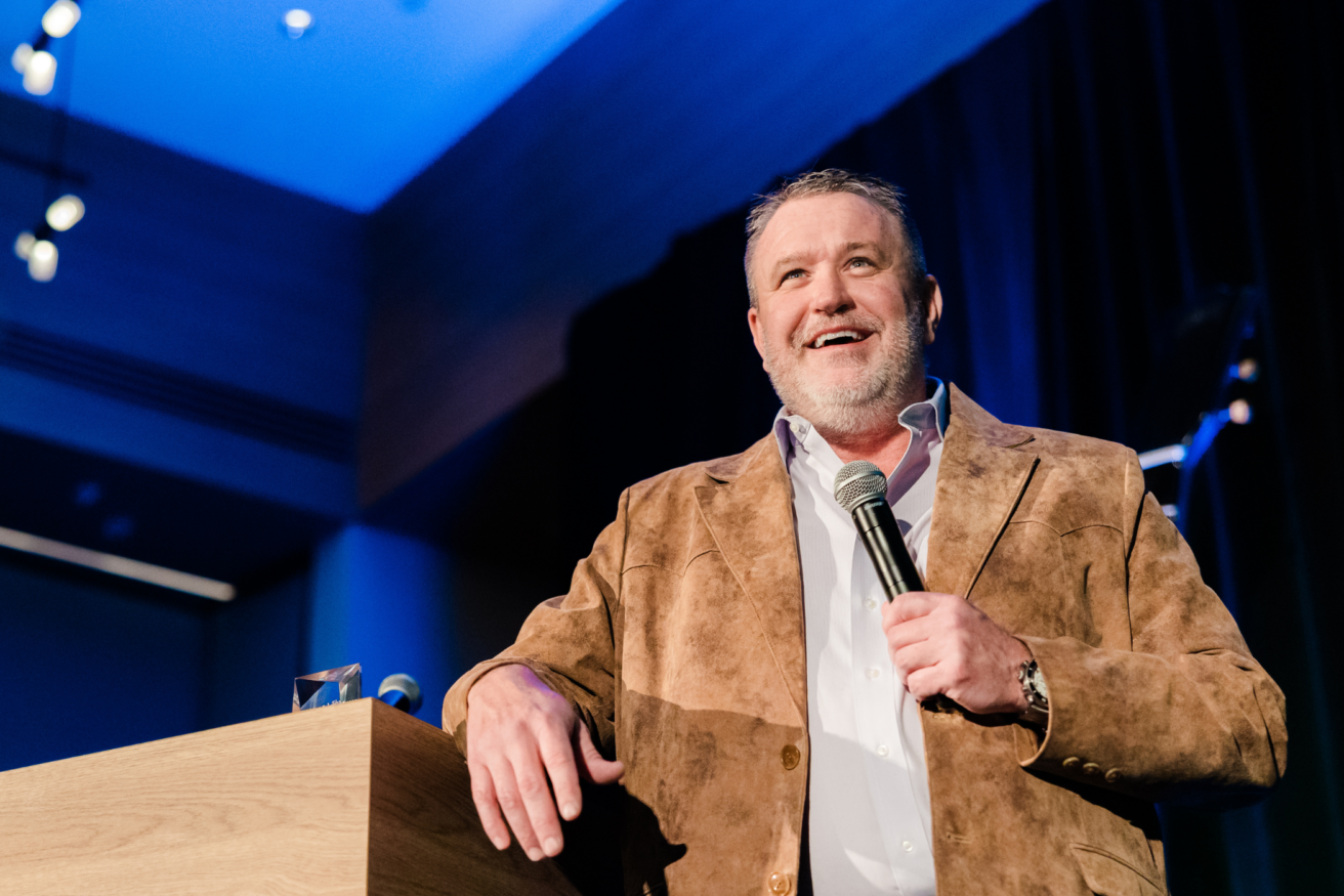 A man with a beard stands at a podium holding a microphone, wearing a brown suede jacket and white shirt, speaking to an audience.