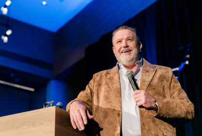 A man with a beard stands at a podium holding a microphone, wearing a brown suede jacket and white shirt, speaking to an audience.