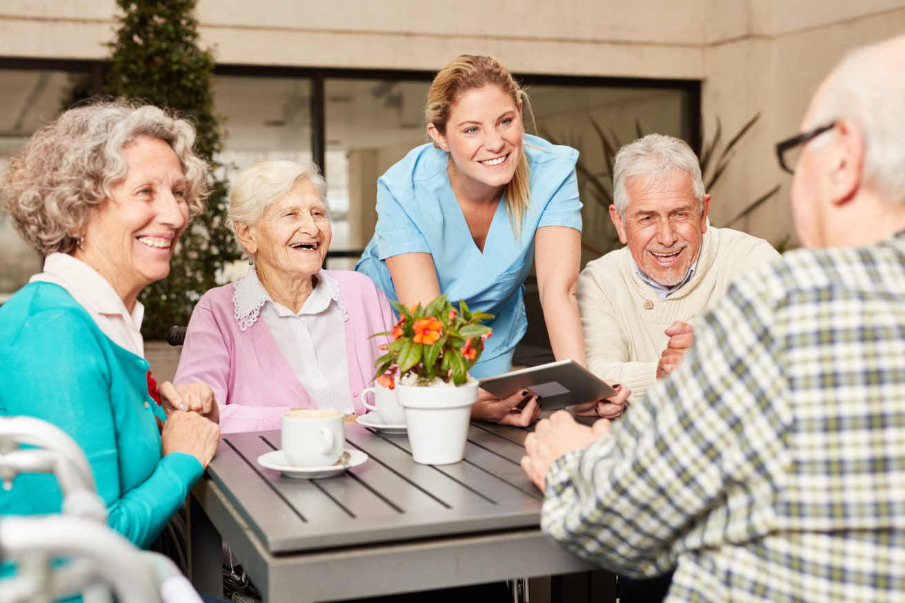 A caregiver in blue scrubs interacts with four elderly people seated around a table outdoors, with plants and a building in the background. A tablet and a potted plant are on the table.