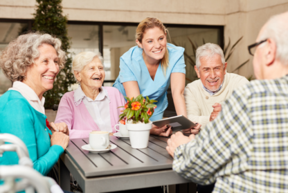 A caregiver in blue scrubs interacts with four elderly people seated around a table outdoors, with plants and a building in the background. A tablet and a potted plant are on the table.