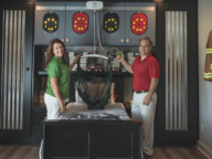 Two adults standing by a child's bed, holding a mobility assistive device. The room has firefighter-themed decorations, including helmets, a firetruck mural, and a hanging firefighter uniform.