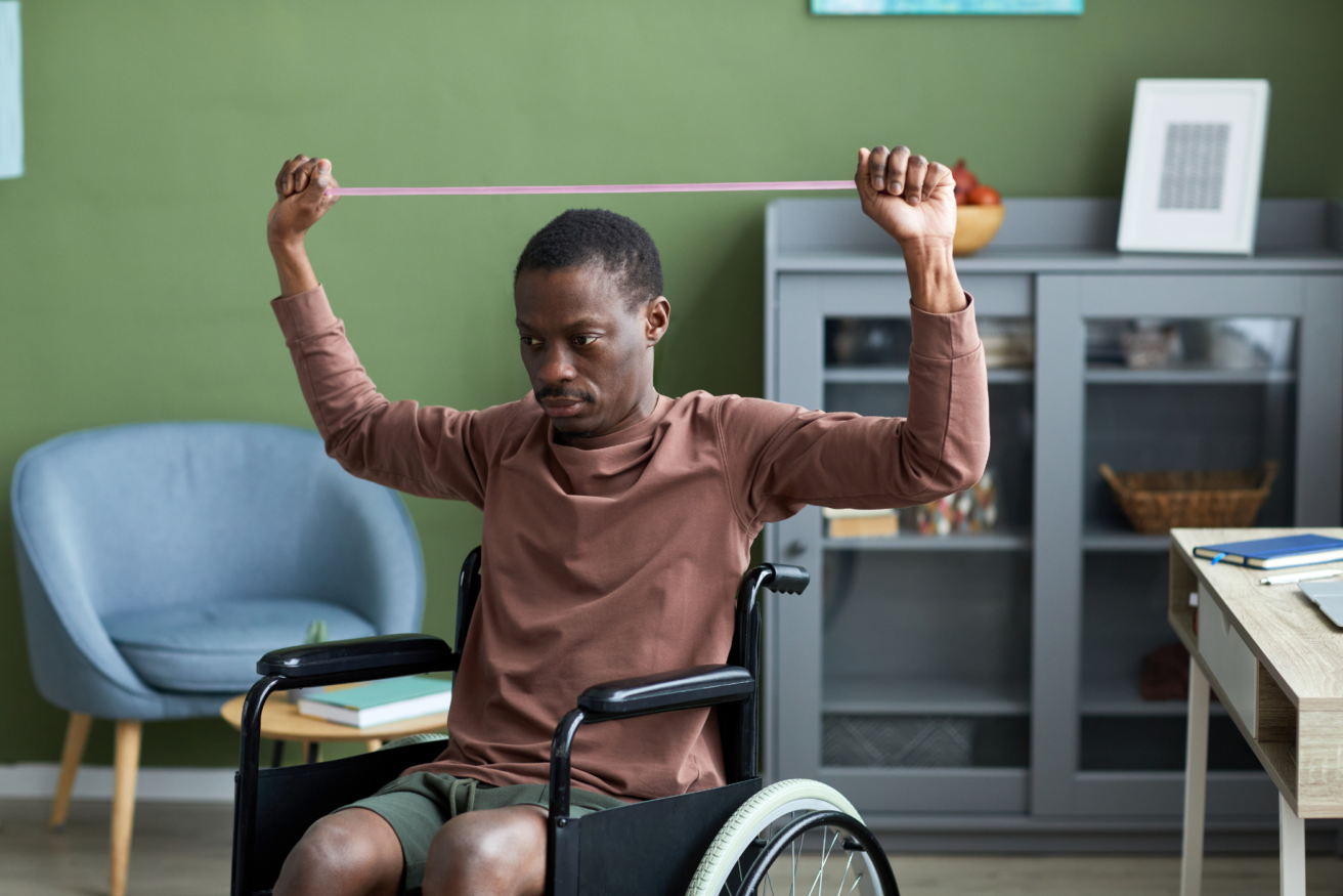 A man in a wheelchair exercises with an elastic resistance band in a room with green walls, a gray cabinet, and a light blue chair.