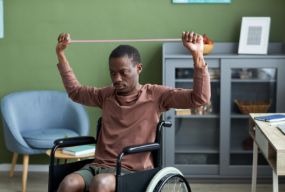 A man in a wheelchair exercises with an elastic resistance band in a room with green walls, a gray cabinet, and a light blue chair.