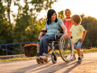 A woman in a wheelchair smiling while two children, a girl and a boy, play and walk alongside her outdoors.