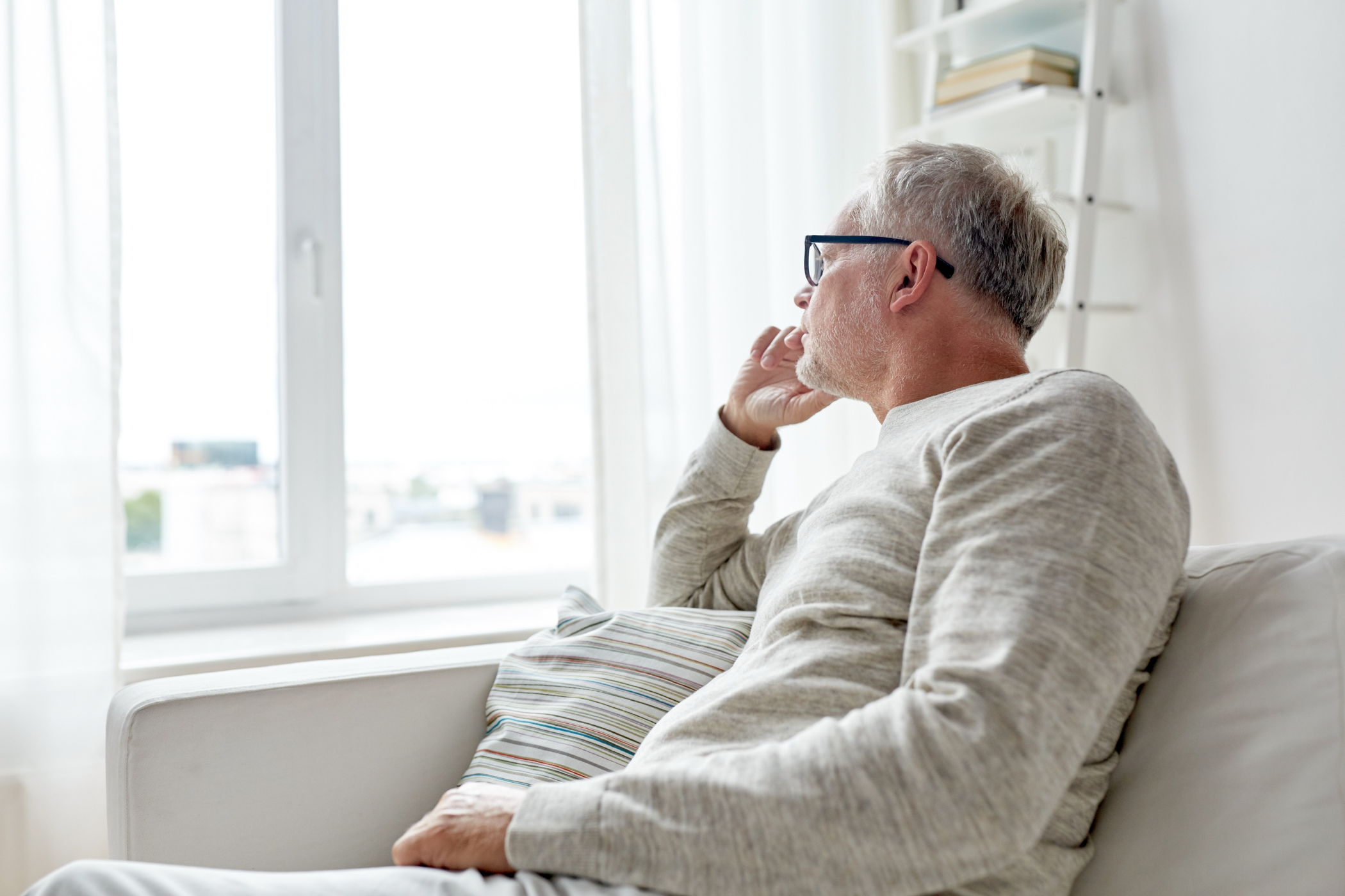 Elderly man with glasses sitting on a couch, looking out a window thoughtfully with a hand on his cheek in a bright room.