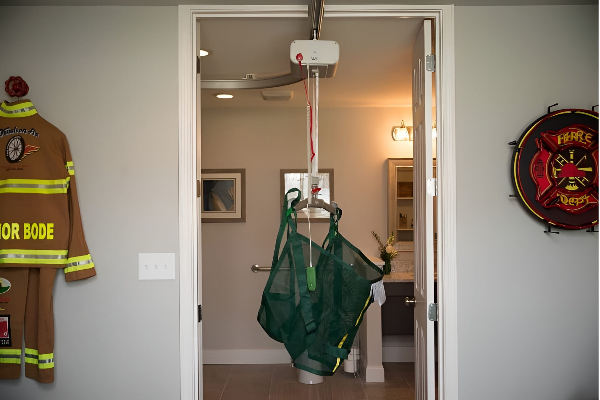 Room with a patient lift system installed on the ceiling, a firefighter's uniform on the wall to the left, and a firefighter insignia on the wall to the right. The room leads into a bathroom.