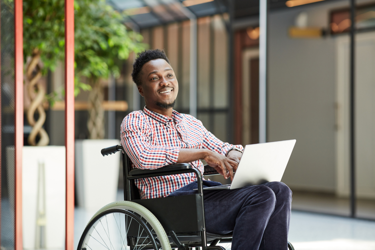 A person in a wheelchair smiles while working on a laptop in a modern indoor setting.