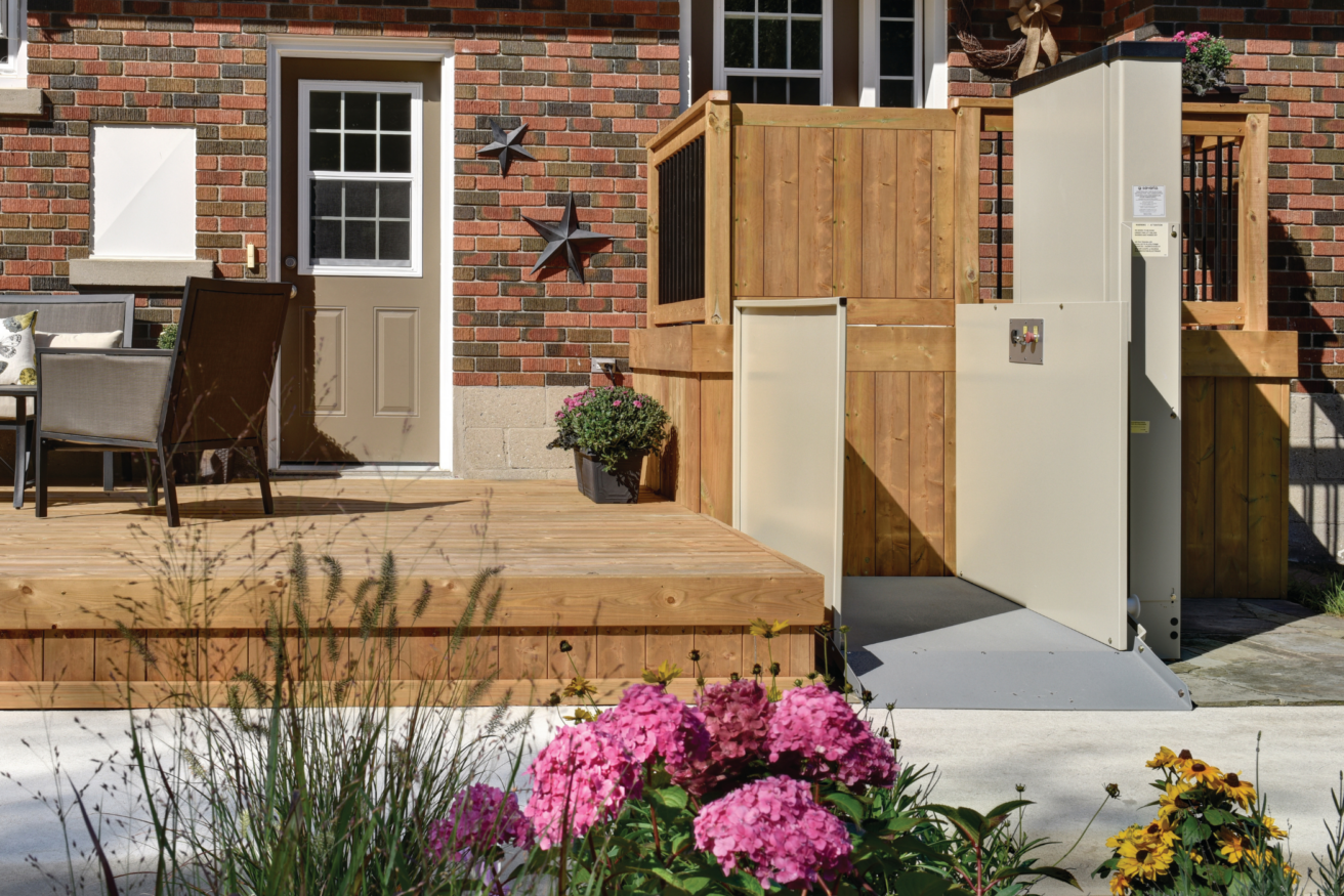 A residential backyard with a brick wall, wooden deck with chairs and table, and a vertical platform lift for accessibility next to a garden with pink and yellow flowers.