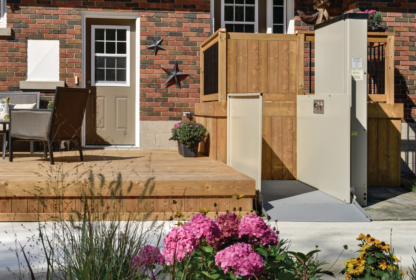 A residential backyard with a brick wall, wooden deck with chairs and table, and a vertical platform lift for accessibility next to a garden with pink and yellow flowers.