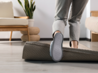 Person tripping over a rolled-up carpet in a modern living room with a wooden chair, a potted plant, and pale wood flooring.