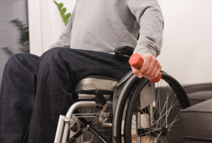 A person in a wheelchair, wearing a grey long-sleeve shirt and jeans, holds a small orange dumbbell.