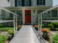 A red door with a wreath is reached via a metal ramp, framed by flowers and bushes, on a white house with large windows and green shutters.