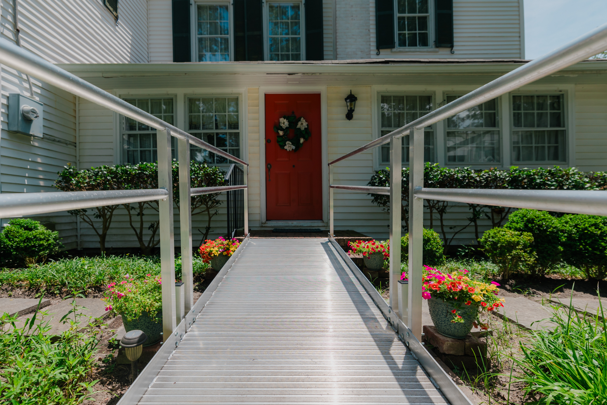 A red door with a wreath is reached via a metal ramp, framed by flowers and bushes, on a white house with large windows and green shutters.