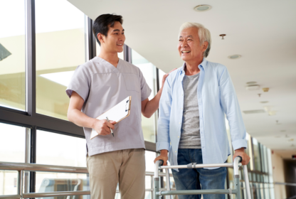 A nurse assists an elderly man using a walker in a corridor, both smiling. The nurse holds a clipboard.