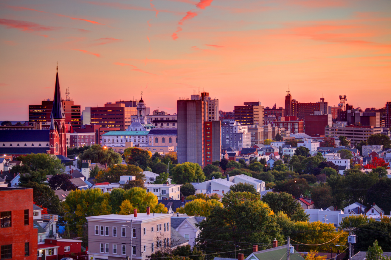 A cityscape view of Portland, Maine at sunset, featuring a mix of older and modern buildings with trees interspersed among the structures.