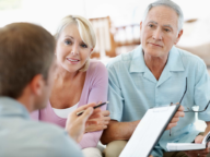 An elderly couple is seated and attentively listening to a man holding paperwork and a pen. The man appears to be explaining or discussing something with them. The couple looks engaged.