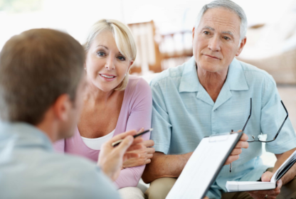 An elderly couple is seated and attentively listening to a man holding paperwork and a pen. The man appears to be explaining or discussing something with them. The couple looks engaged.