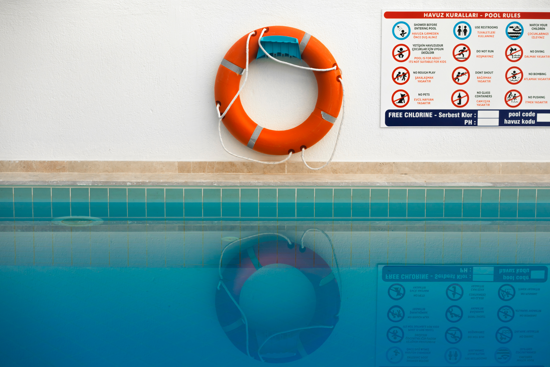 A life preserver hangs on a wall next to a pool rules sign above a swimming pool.