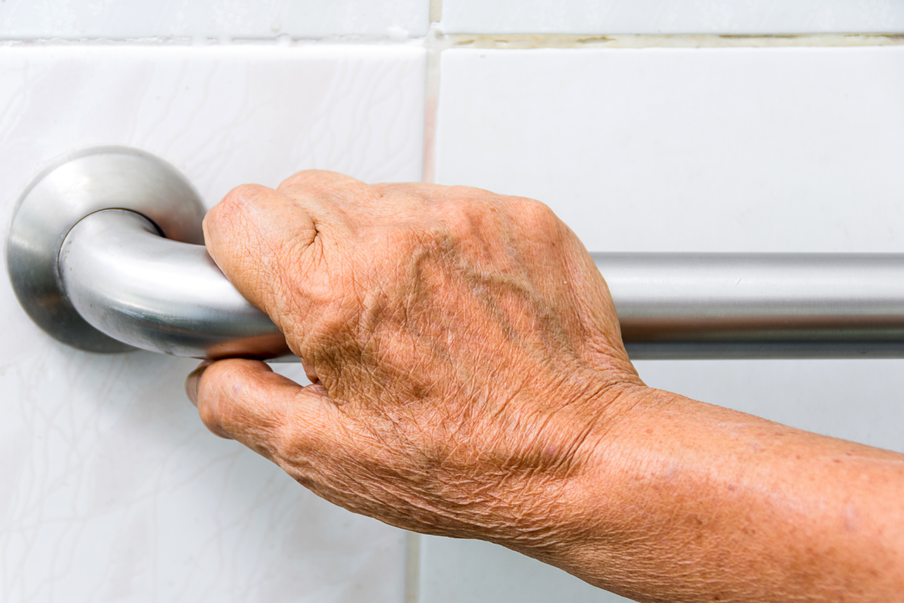 An elderly hand gripping a metal safety bar mounted on white tiled wall.