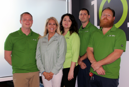 A group of five people, four men and one woman, stand indoors wearing green shirts. They are positioned in front of a wall with the number "101" visible in the background.