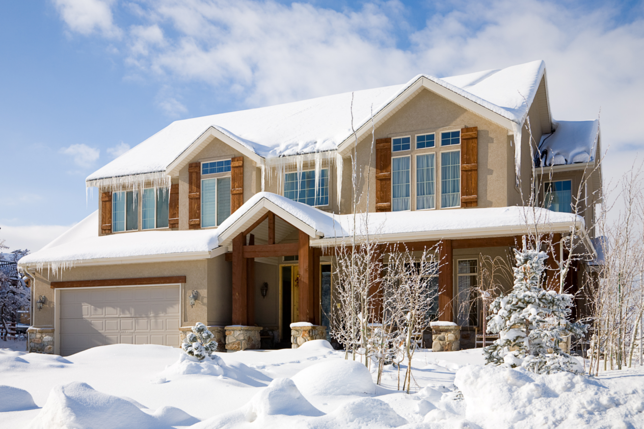 A two-story house with wooden accents is covered in snow and surrounded by a snowy landscape under a clear blue sky.