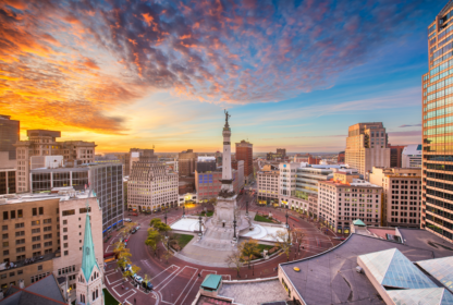 Aerial view of a city square at sunset with a central monument, several buildings surrounding the square, and a vibrant, colorful sky in the background.