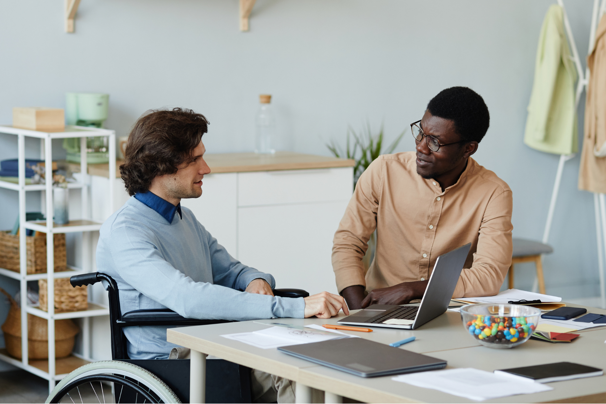 Two men sitting at a table in an office, one in a wheelchair, having a discussion and looking at a laptop. Office supplies and a bowl of colorful candies are on the table. Shelves are in the background.