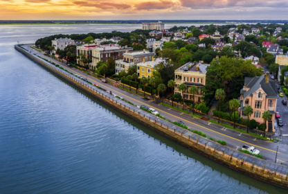 Aerial view of a waterfront neighborhood with large houses lined along a tree-lined street beside a body of water during sunset.