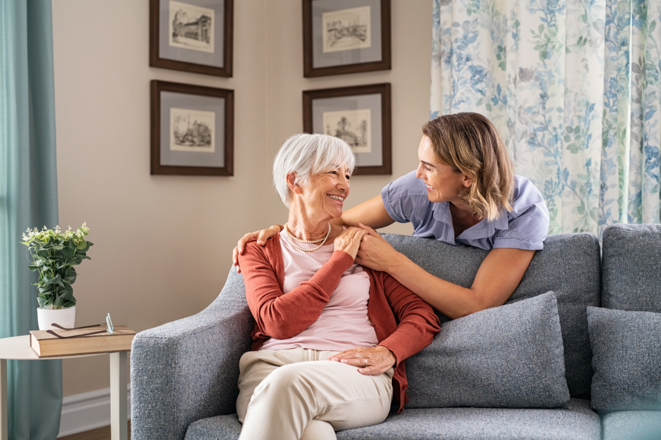 An elderly woman sits on a sofa, smiling and holding hands with a younger woman who is leaning over the back of the sofa. Framed pictures and floral curtains are visible in the background.