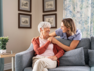 An elderly woman sits on a sofa, smiling and holding hands with a younger woman who is leaning over the back of the sofa. Framed pictures and floral curtains are visible in the background.