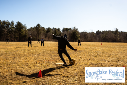 A group of people playing kickball on a grassy field with a "Snowflake Festival Charity Kickball Tournament" sign displayed in the corner. Weather appears cold, and players are dressed in warm clothing.