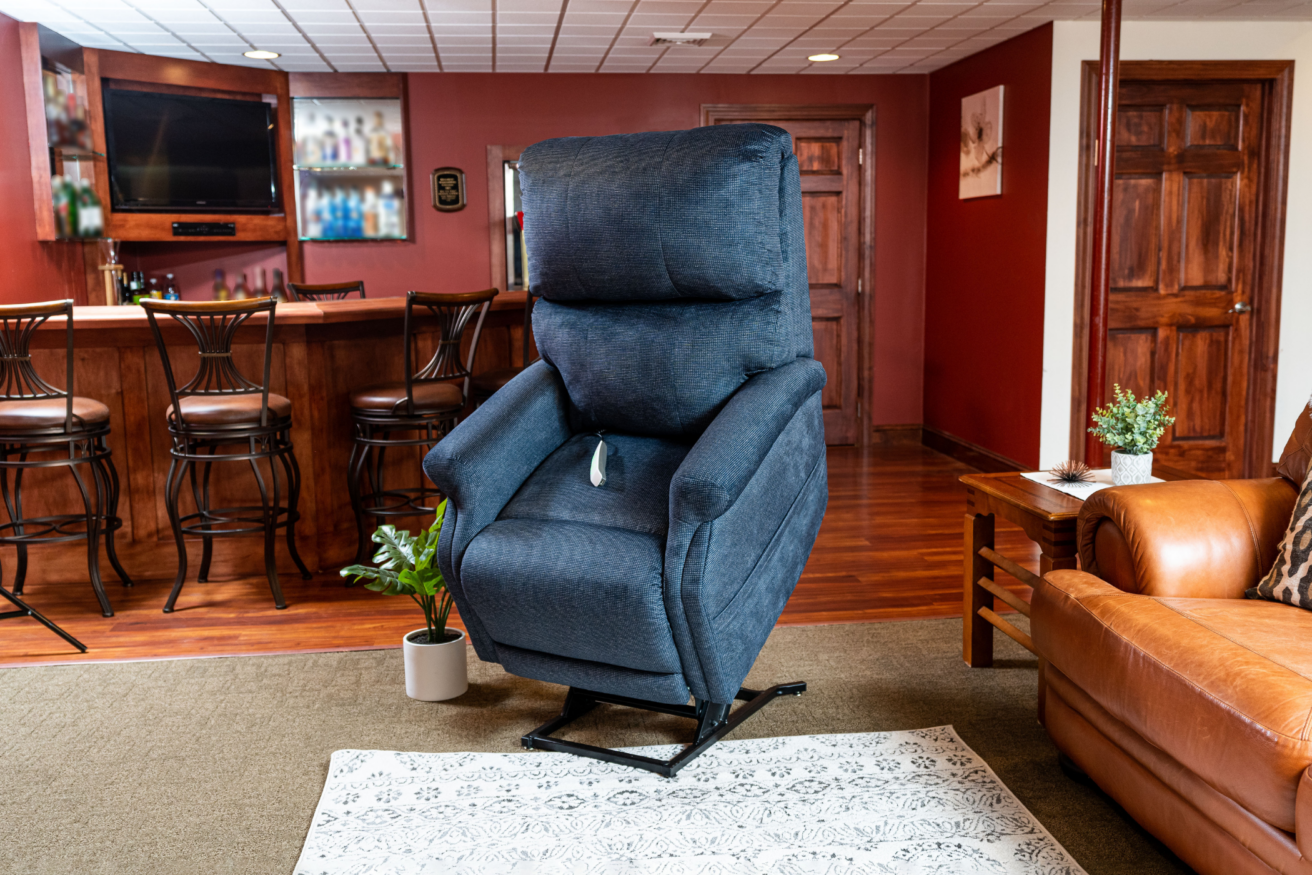 A blue reclining lift chair is positioned in a living room near a leather sofa and wood-paneled bar area. A white patterned rug lies on the floor, and a small potted plant sits beside the chair.