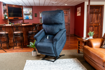 A blue reclining lift chair is positioned in a living room near a leather sofa and wood-paneled bar area. A white patterned rug lies on the floor, and a small potted plant sits beside the chair.