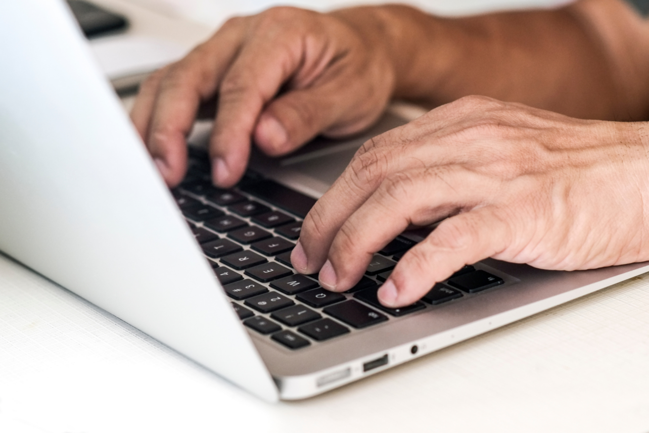 Close-up of hands typing on a laptop keyboard. The laptop is open on a light-colored surface.